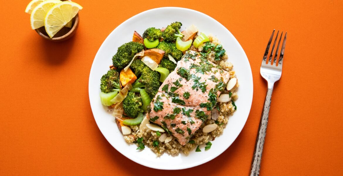 A plate of herb-roasted salmon served over quinoa and topped with sliced almonds, accompanied by roasted vegetables including broccoli, celery, onion, and sweet potato.