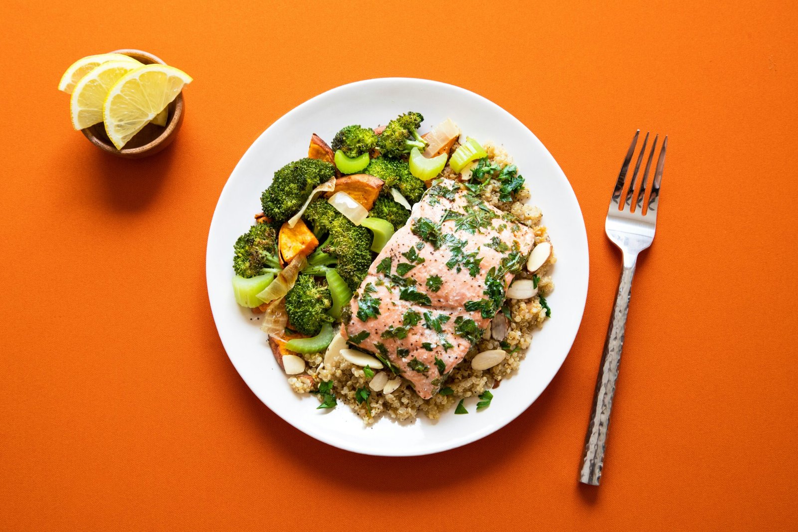 A plate of herb-roasted salmon served over quinoa and topped with sliced almonds, accompanied by roasted vegetables including broccoli, celery, onion, and sweet potato.